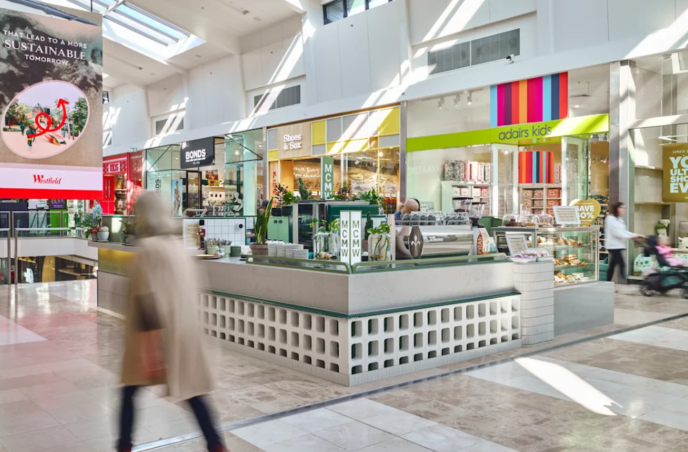 Interior of a shopping mall with various retail stores, a coffee kiosk, and shoppers walking by. Bright natural light filters through skylights.