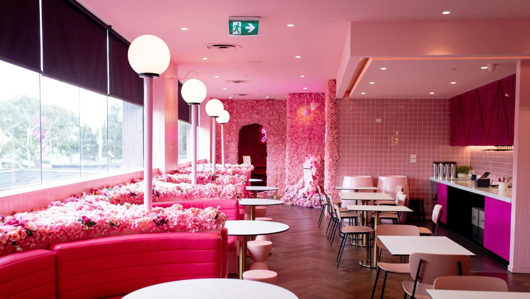 Interior of a cafe or restaurant with pink decor, floral accents, pink booths, white tables, and pink tiles, illuminated by round ceiling lights.