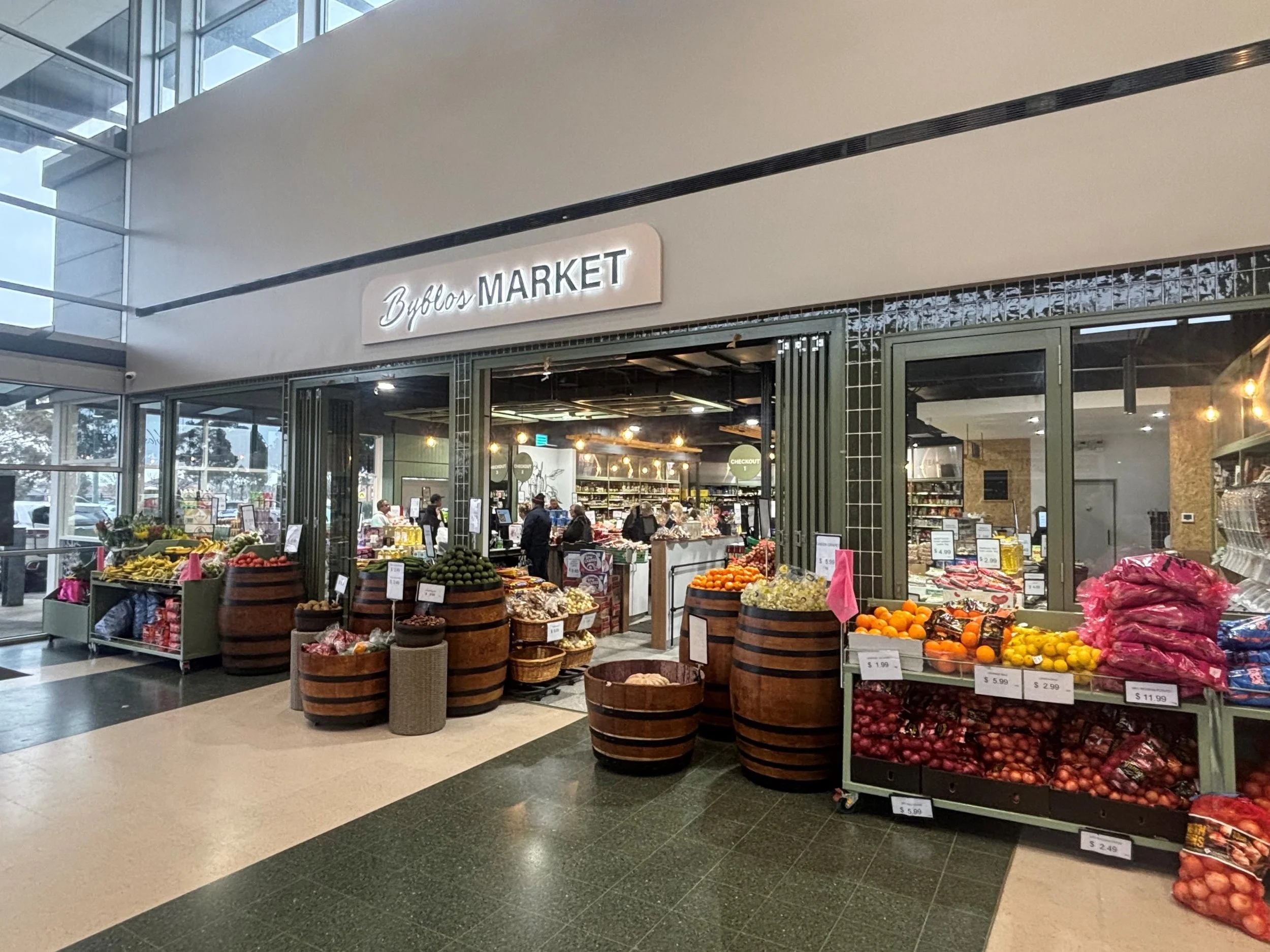Entrance to a grocery store called Byblos Market with fresh produce displayed outside, including oranges, apples, and vegetables, inside customers shopping.