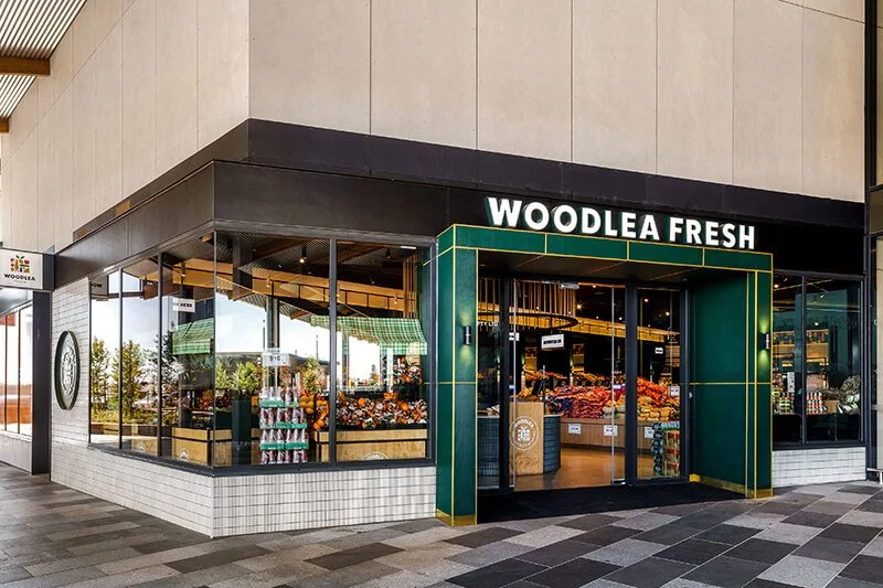 Exterior view of Woodlea Fresh, a grocery store with large glass windows, showcasing fresh produce inside. The store has a green and gold entrance canopy and is located in a modern shopping area.