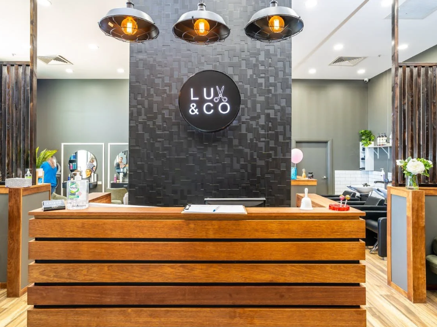 Reception desk at Lu & Co salon with black textured wall, illuminated sign, and overhead industrial lights.