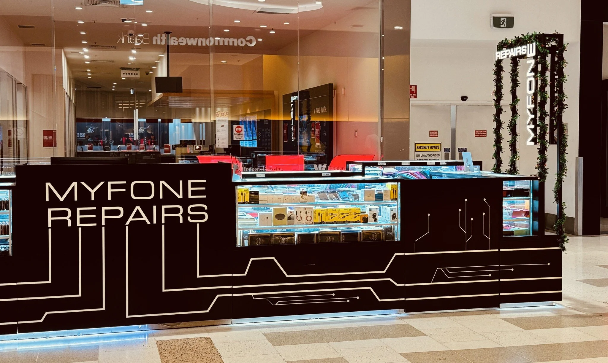 A black electronic repair kiosk labeled "MYFONE REPAIRS" with circuitry design lines, glass display cases containing phone accessories and repair tools, located inside a shopping mall. Background includes glass walls and signage.