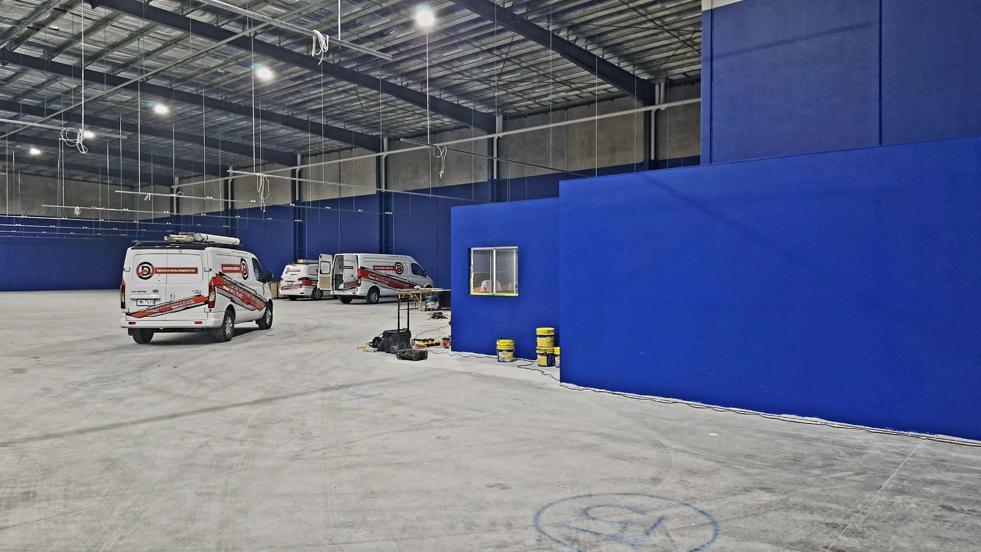 Inside a large industrial building under construction with blue walls, multiple work vans, construction tools, and equipment on the floor.