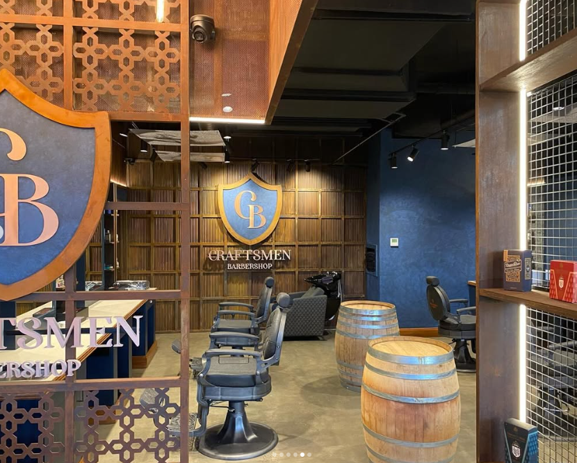 Interior of a barbershop with leather barber chairs, a counter, and wooden barrel tables. The walls feature a large sign with the initials 'CB' inside a shield, and the decor includes wooden accents and wood paneling.