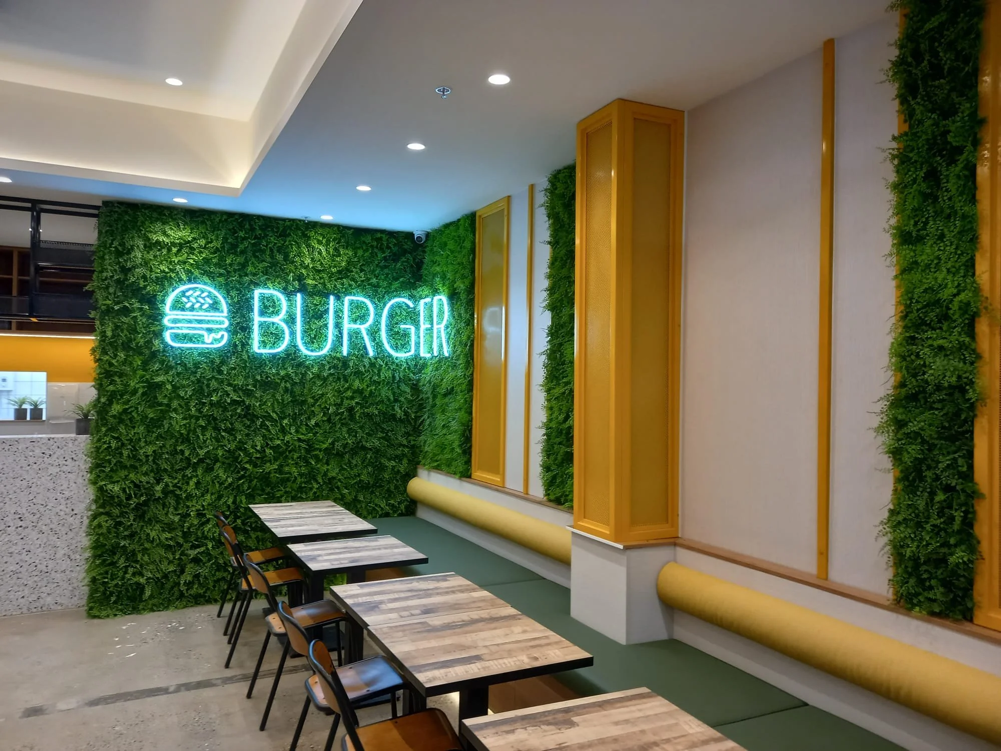 Interior of a burger restaurant with a green leafy wall, neon sign reading "BURGER" and a burger icon, wooden tables and chairs, and beige walls with yellow accents.