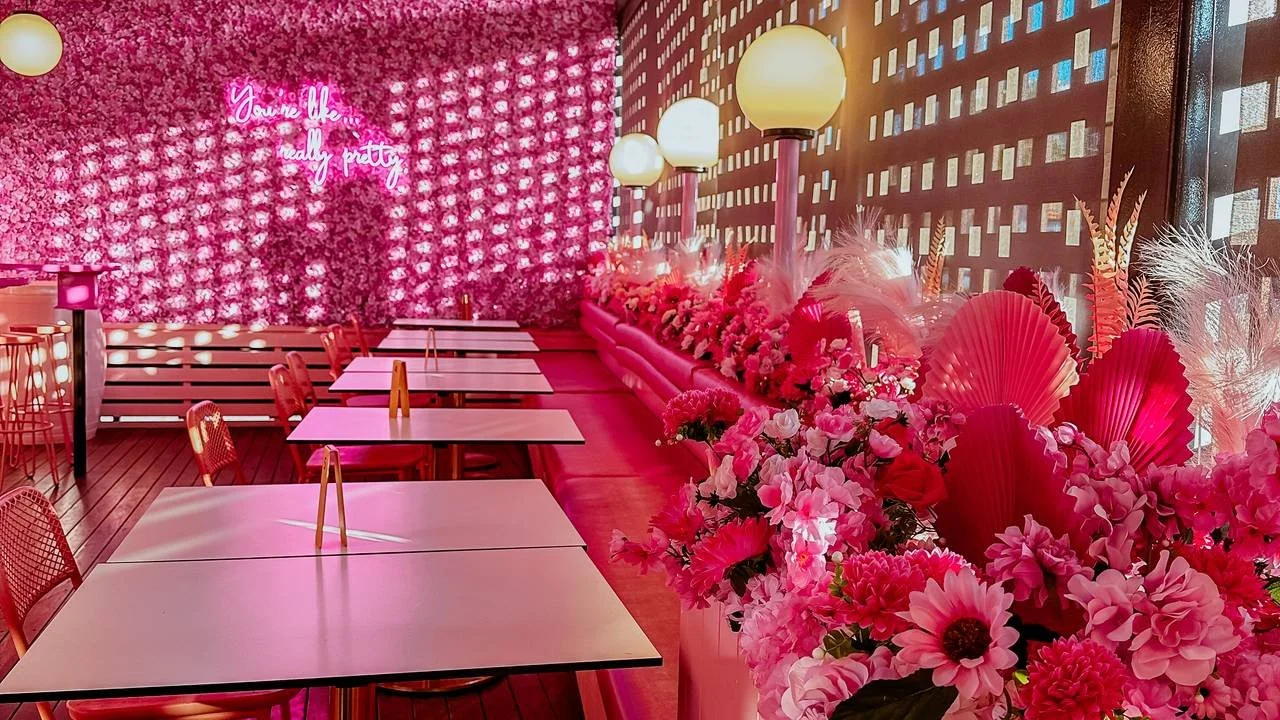 A restaurant or cafe with pink and floral decor, pink tables, wooden chairs, a floral wall with neon sign, and pink lighting.