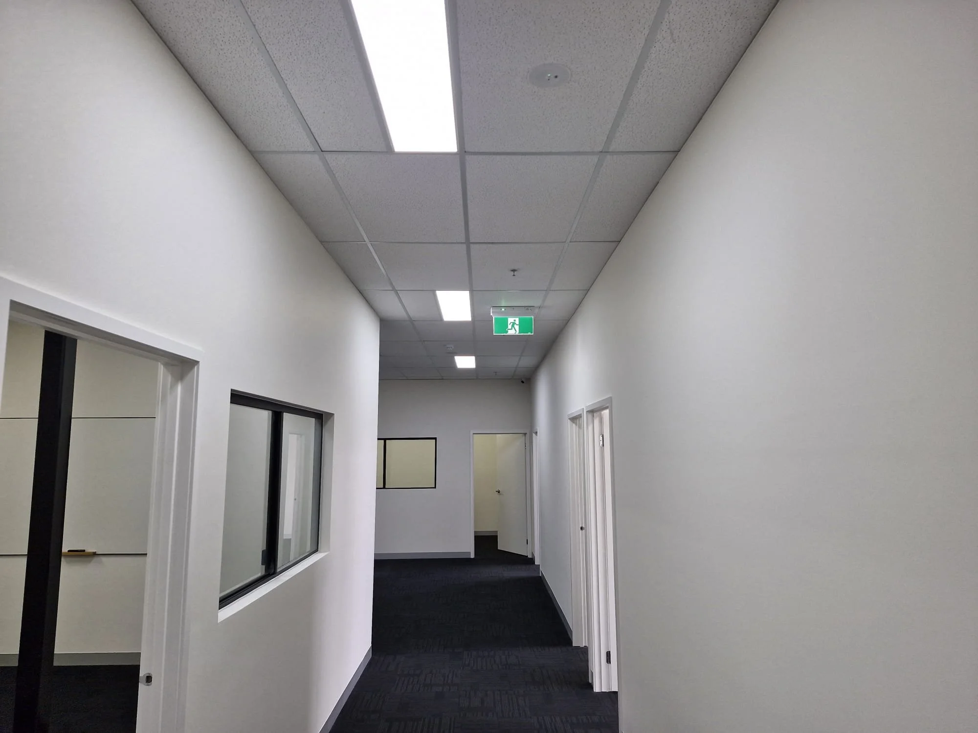 Empty hallway with white walls, ceiling lights, windows on the left, and an exit sign hanging from the ceiling.