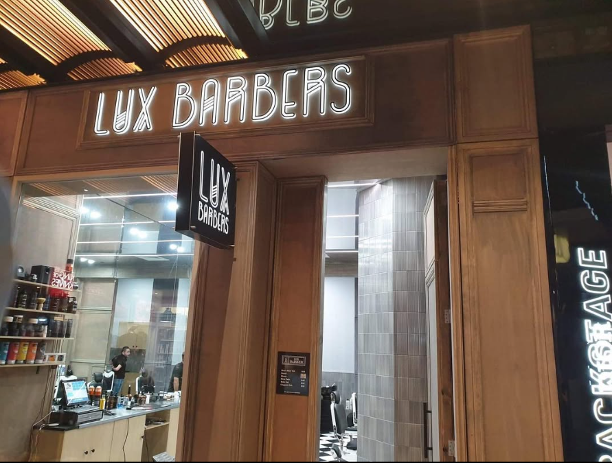 Entrance to Lux Barbers with wood-paneled walls, illuminated sign, and view into barber shop interior with shelves stocked with hair products and barber chairs.