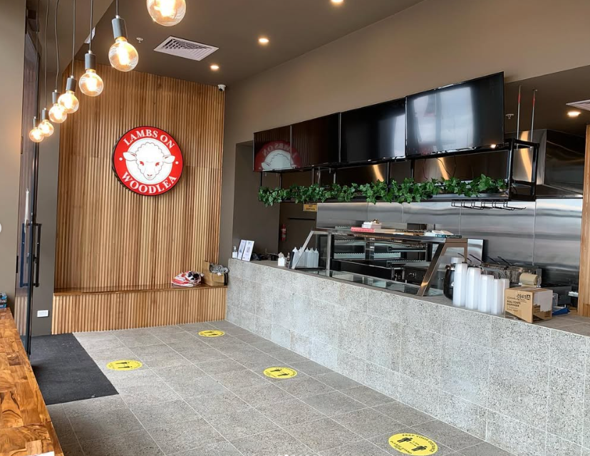Interior of a restaurant or food establishment with a wood-paneled wall, a Lambs & Woodlea sign, and a counter area with multiple TV screens, greenery, and drink cups.