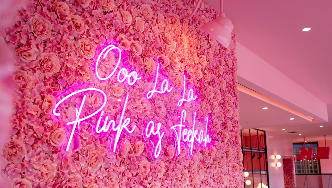 Pink floral wall with neon sign that reads "Ooo La La Pink ay Stekah" in cursive lettering.