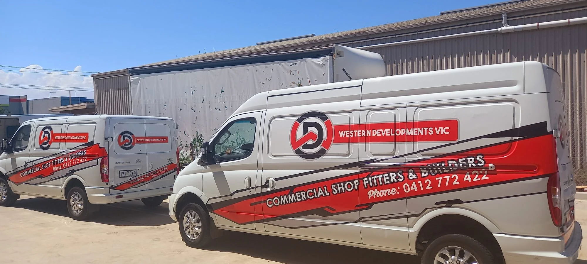 Two white commercial vans with red and black branding for Western Developments VIC, parked outdoors on a sunny day, with a large industrial building in the background.