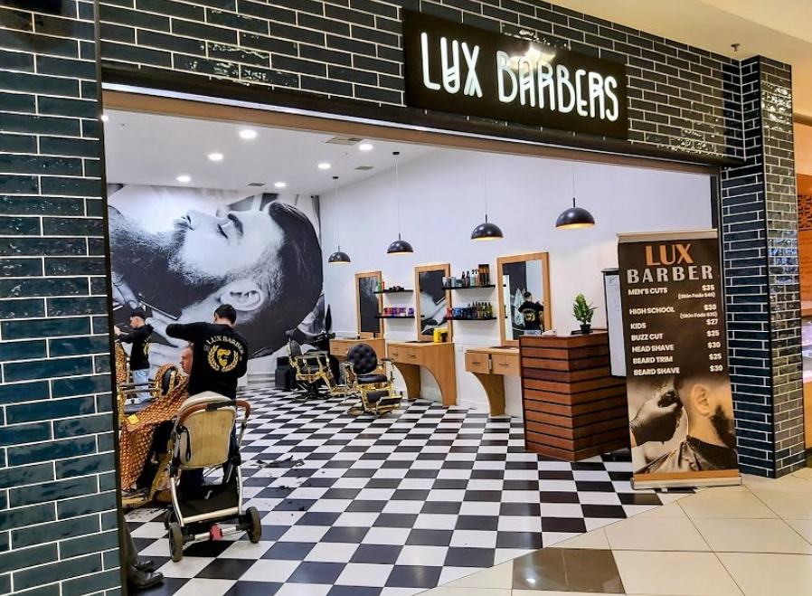 Inside a barbershop named Lux Barbers with black brick walls, checkered black and white floor, and modern decor. Clients are getting haircuts, and there's a large black-and-white mural of a woman on the back wall. The shop has barber chairs, mirrors,