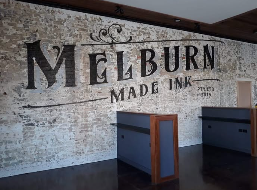 Interior of a building with a white brick wall featuring large black text that reads "Melburn Made Ink" with decorative elements, and two wooden-topped gray counters.