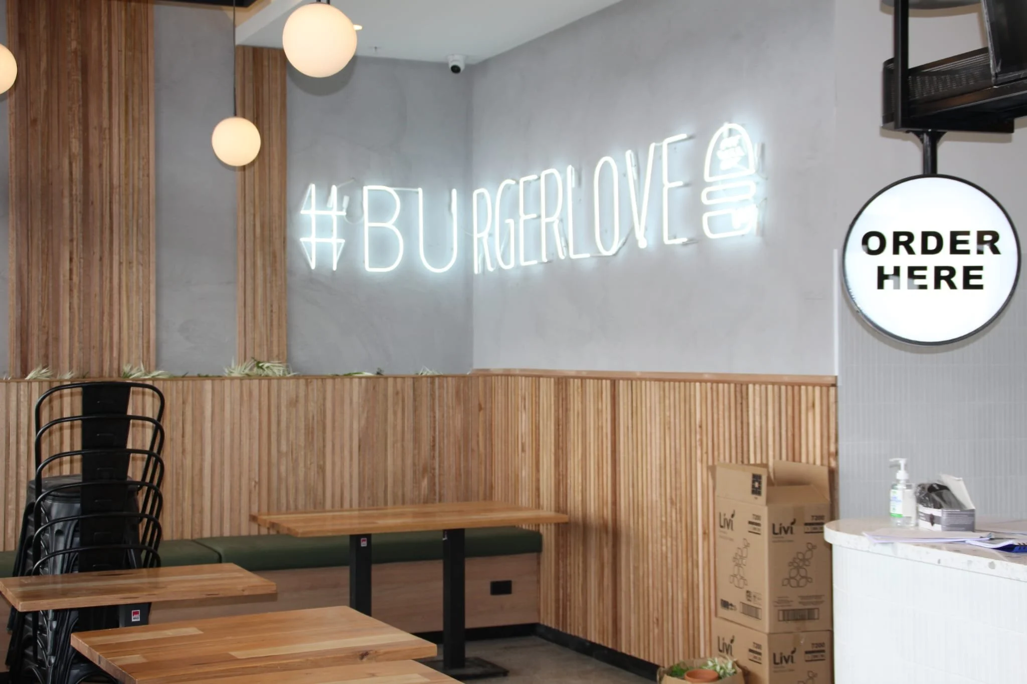 Interior view of a burger restaurant with stacked chairs, wooden tables, a wall with wood paneling, a neon sign that reads '#BURGERLOVE,' and a round sign that says 'ORDER HERE' near the counter.