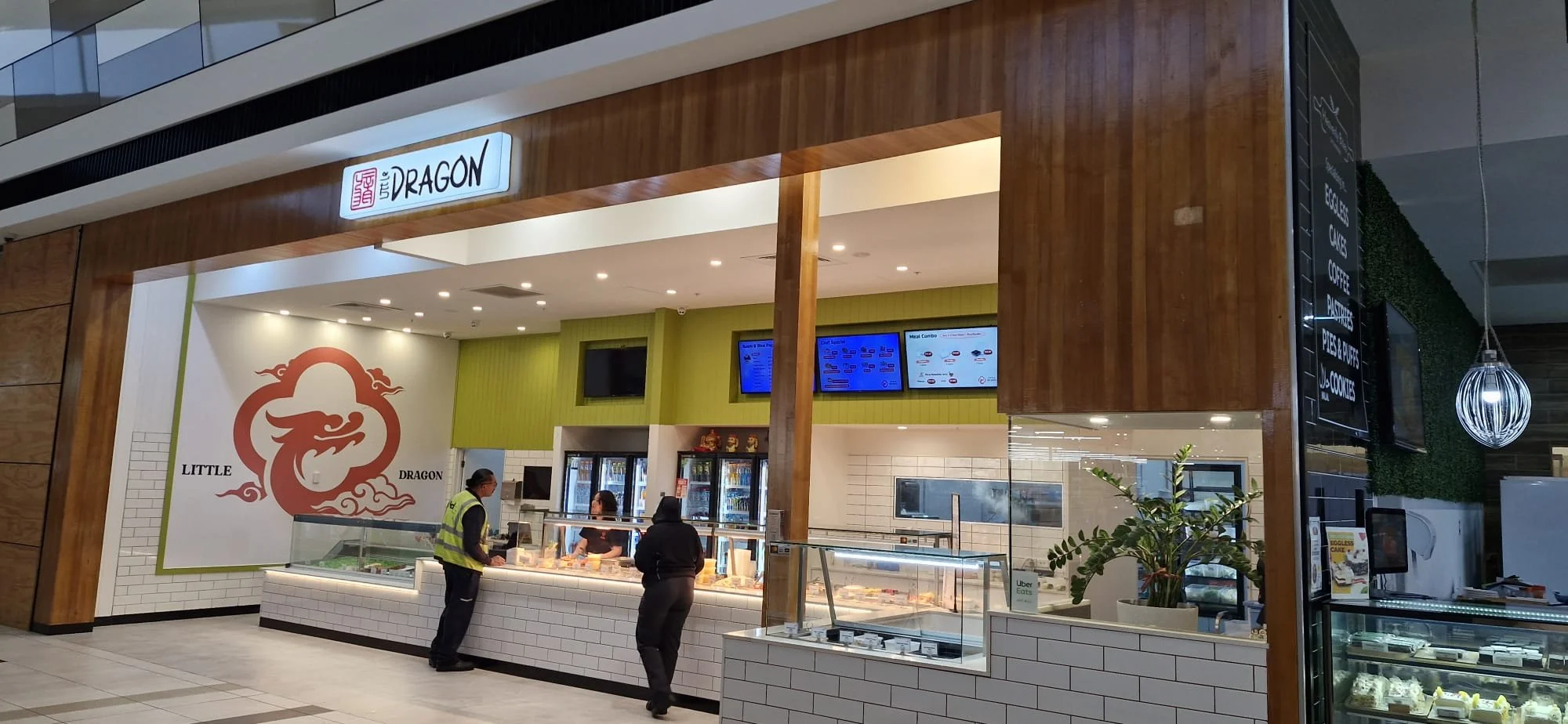 A food stall named Little Dragon inside a shopping mall with a wooden frame, white brick counter, and digital menu screens overhead. There are two customers ordering, and a staff member behind the counter. The stall has a large logo of a stylized red