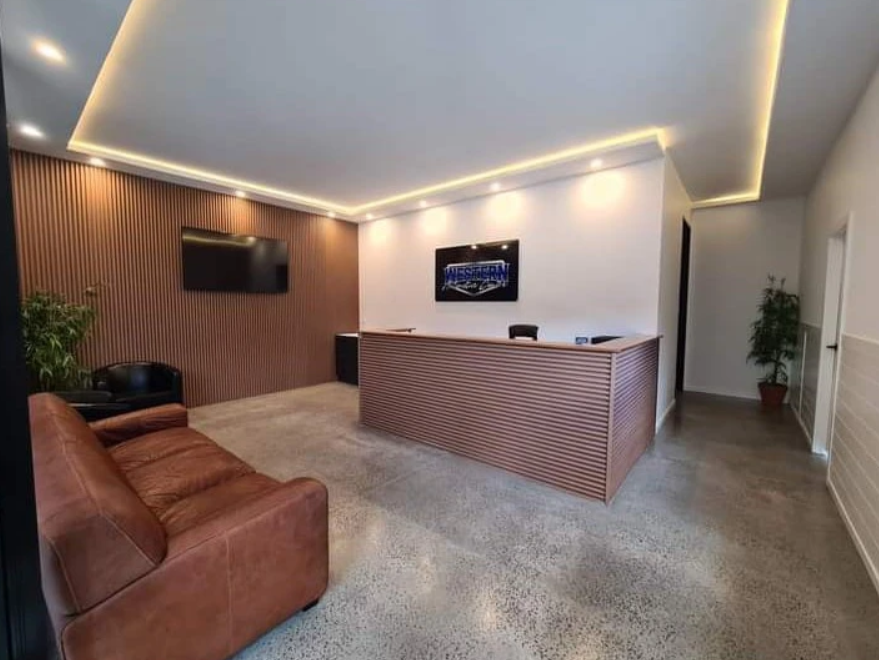 Modern office reception area with a wooden front desk, a leather couch, a TV on the wooden accent wall, potted plants, and a flat-screen monitor.