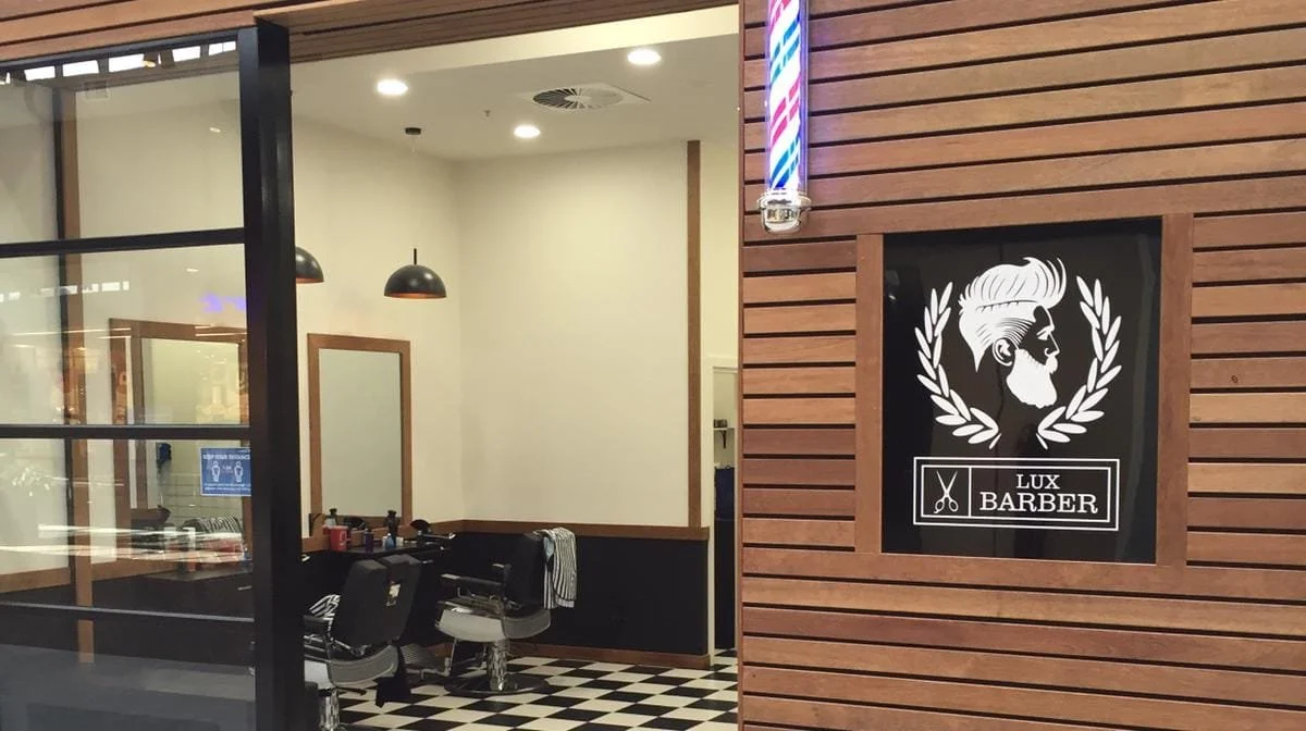 Interior of a barbershop with a black and white checkered floor, a large mirror, barbershop chairs, and a barber sign on the wooden wall outside.