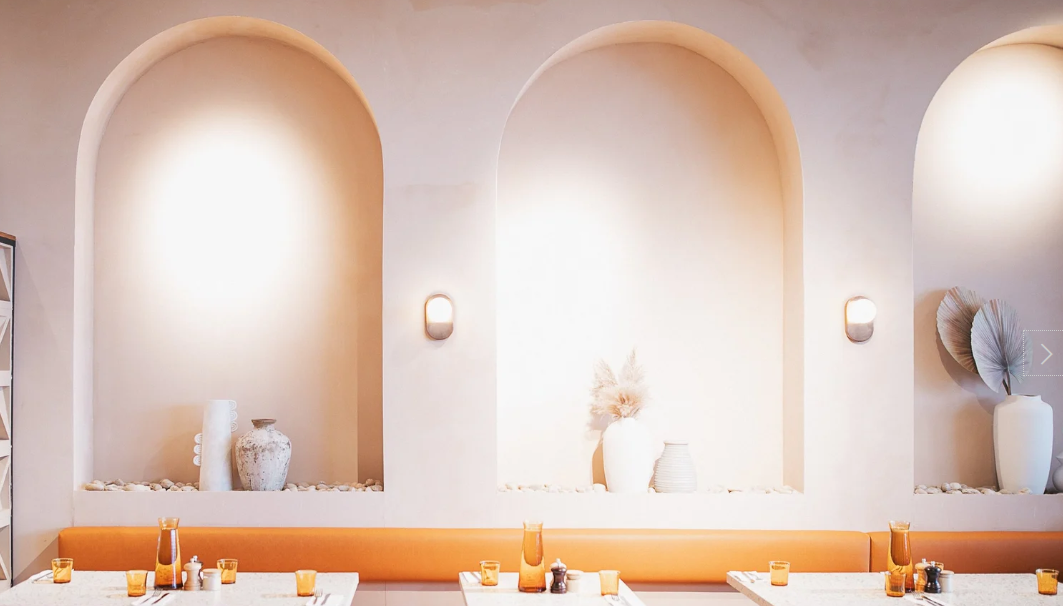 Interior of a modern restaurant with arched wall niches decorated with vases and dried plants, warm lighting, and a long seating area with tables set with cups and candles.