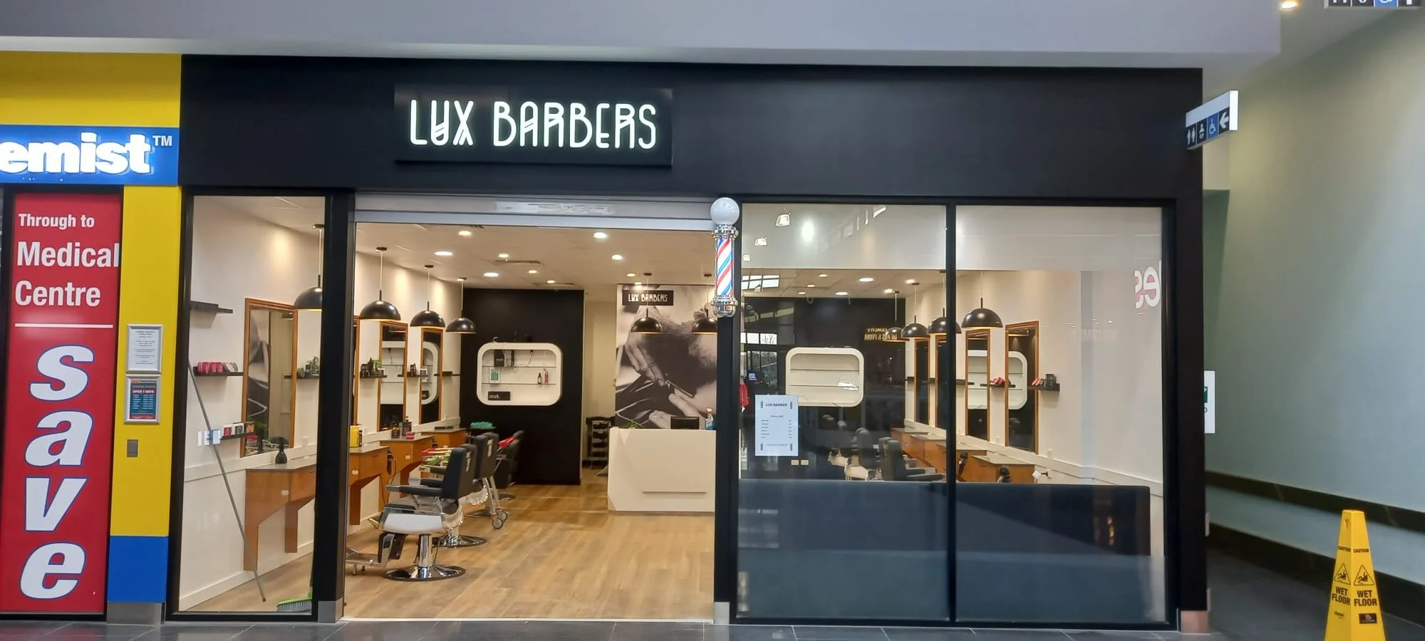 Exterior view of a barbershop named Lux Barbers with black signage, large glass windows, and a barber pole hanging outside. Inside, there are barber chairs, mirrors, and staff preparing for haircuts.