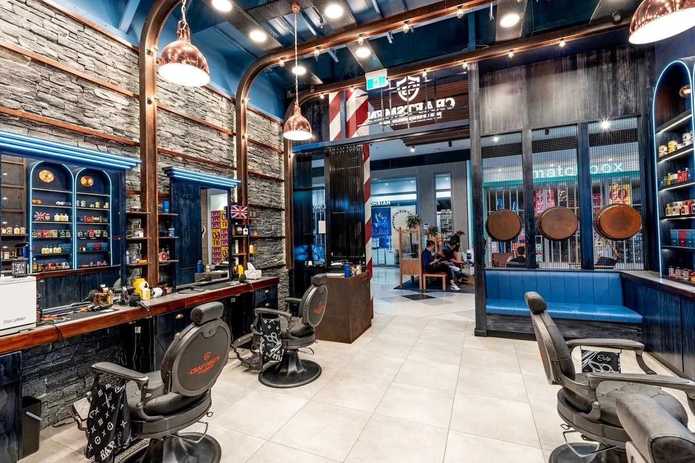 Interior of a modern barber shop with two barber chairs, a wall of shelves with grooming products, and a seating area with people waiting.
