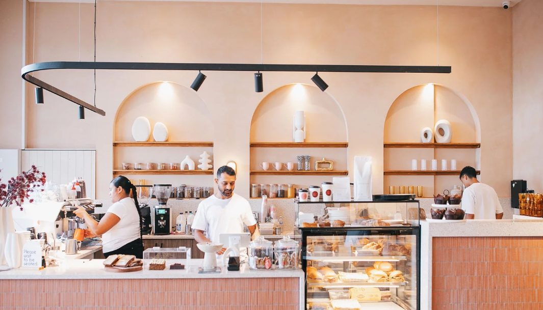 Interior of a modern bakery or coffee shop with staff preparing drinks and food behind the counter, display case with baked goods, minimalist decor with shelves of ceramics and a warm color scheme.