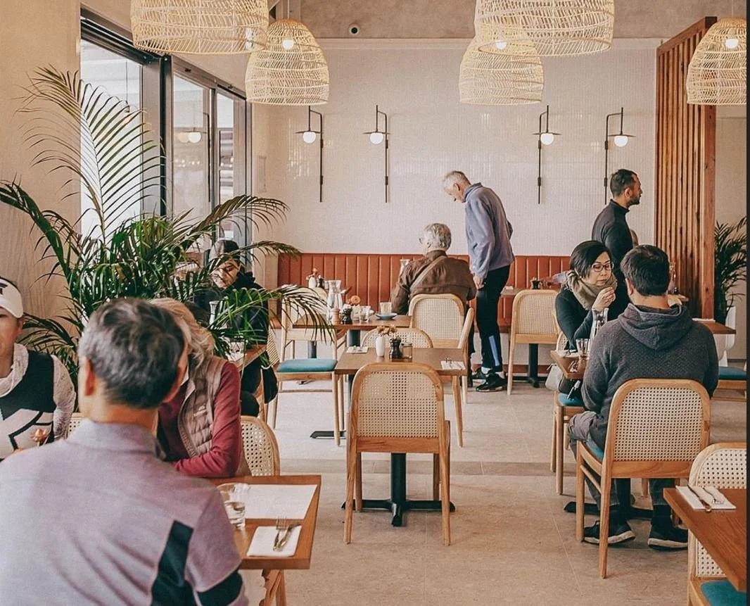 A cozy restaurant interior with several people dining, wooden furniture, large window, plants, and hanging wicker light fixtures.