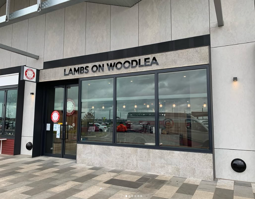 Exterior of a restaurant called Lambs on Woodlea with large glass windows and a door, located in a modern building with a sidewalk in front. The restaurant's name is displayed above the windows.