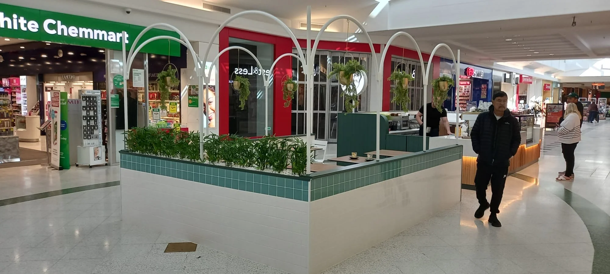 A mall corridor with a small cafe stand with hanging plants and customers inside and nearby.