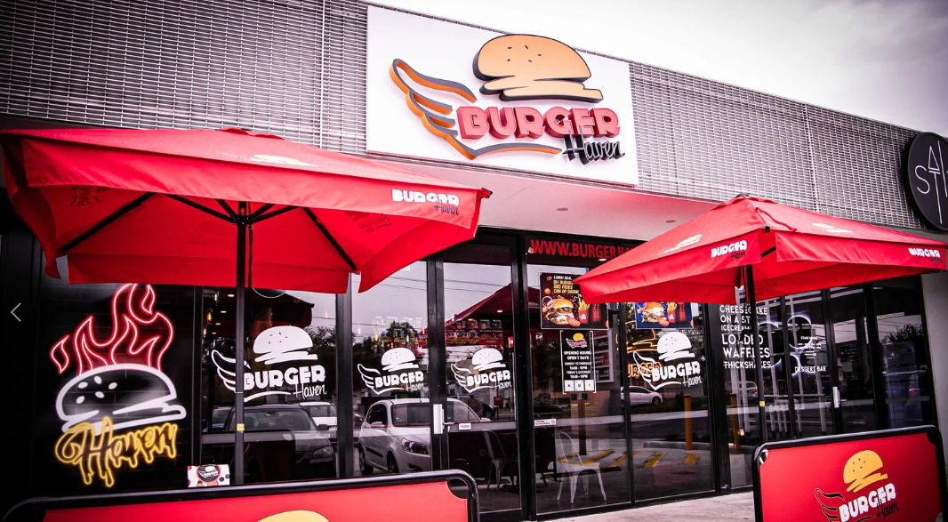 Exterior view of a Burger Haven restaurant with red umbrellas and neon signs, including a hamburger with flames and the Burger Haven logo.