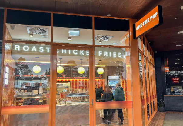 View of a restaurant window with signs for roast chicken and fries, showing interior seating and customers inside.