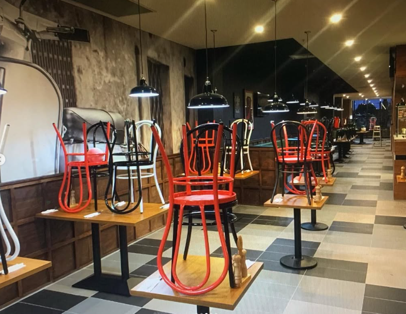 Upside-down chairs stacked on tables inside a dimly lit restaurant or cafe with pendant lights and checkered flooring.
