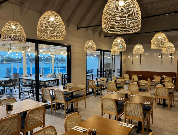 Interior of a modern restaurant with wooden tables, rattan chairs, large woven pendant lights, and floor-to-ceiling windows showing an outdoor dining area at sunset.