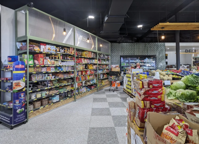 A grocery store aisle with shelves stocked with various packaged food items, fresh vegetables like lettuce, and a refrigerated section in the background.