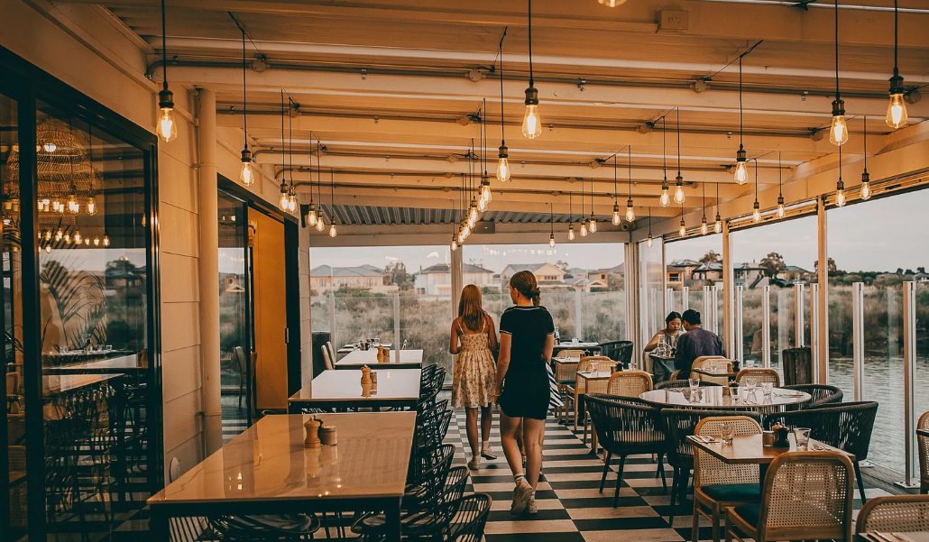 Interior of a restaurant with large glass windows overlooking a river and houses in the distance. The space has hanging Edison bulbs, wooden furniture, and a black-and-white checkered floor. Two women walk through the restaurant, while a few groups o