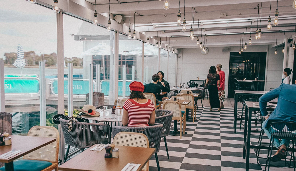 Interior of a modern cafe with checkered black and white floor, string lights, large windows overlooking water, and several people seated at tables, some socializing.