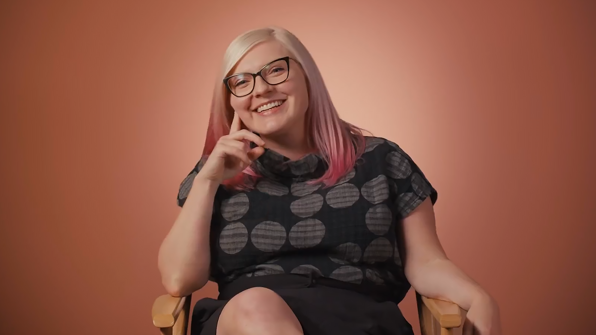 Kim Sutton sits in a directors chair on a film set in front of a peach colored background.