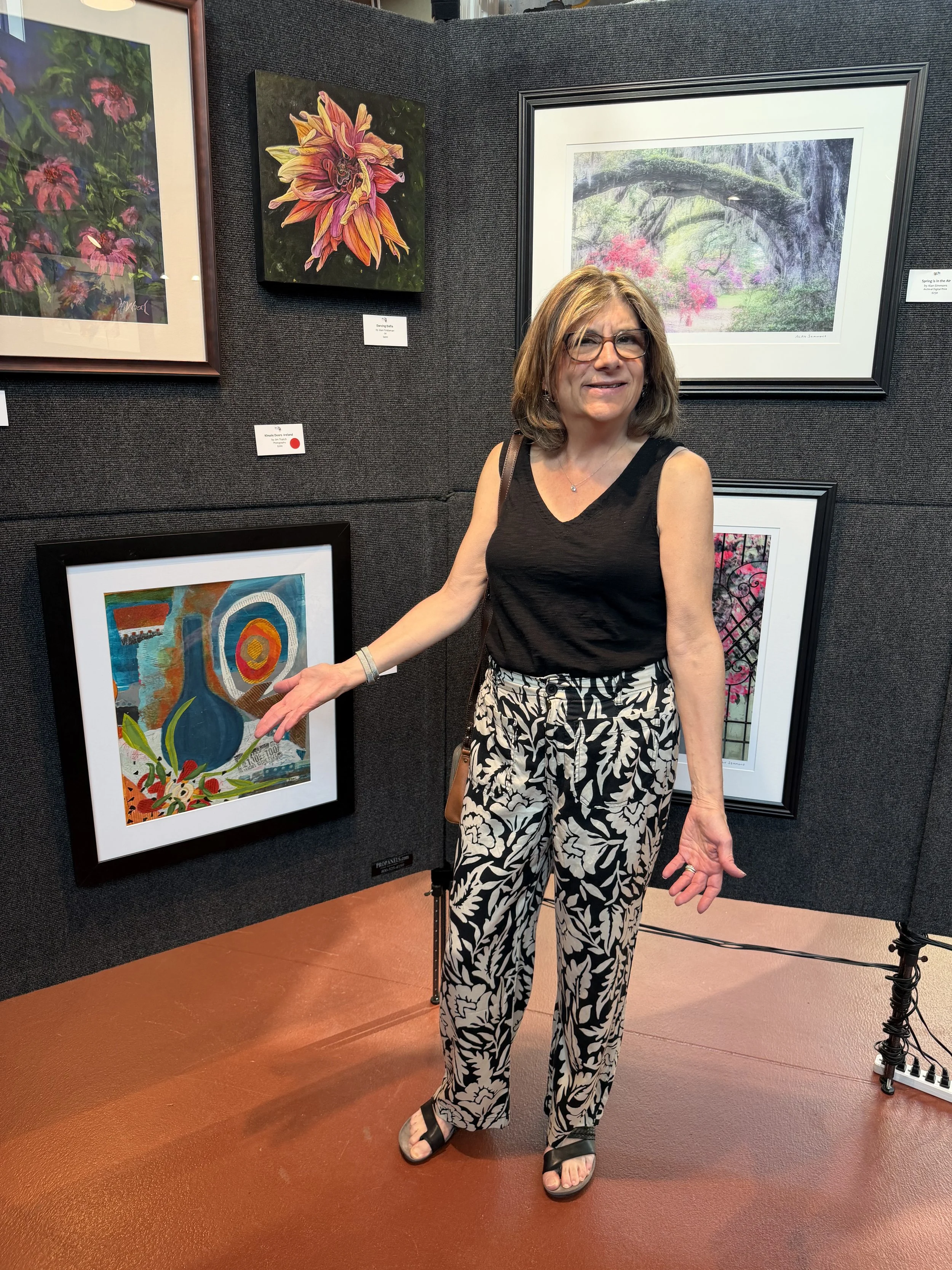 A woman with short brown hair, glasses, wearing a black sleeveless top and patterned black-and-white pants, stands in an art gallery. She is smiling and looking at the camera while pointing at a framed colorful abstract painting on the wall behind her.