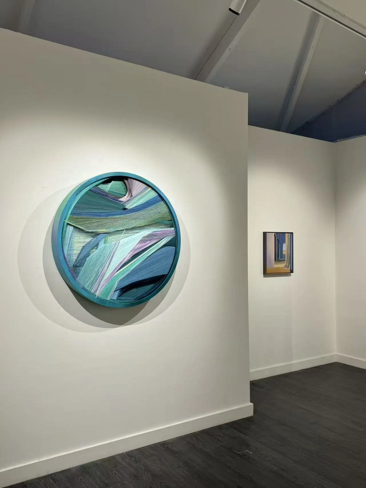 An art gallery with white walls displaying abstract paintings. A large, circular, colorful abstract artwork is mounted on the wall in the foreground, and two smaller paintings are visible on the adjacent wall.