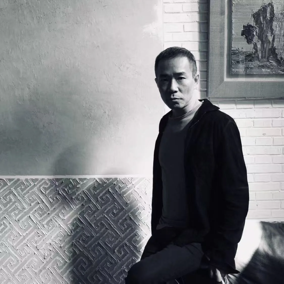 A black and white photo of a man sitting indoors against a textured wall, with artwork in a frame on the wall behind him.
