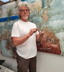A man with gray curly hair and glasses wearing a white t-shirt stands in front of a mural wall with maps and illustrations, holding a small object in his hand.