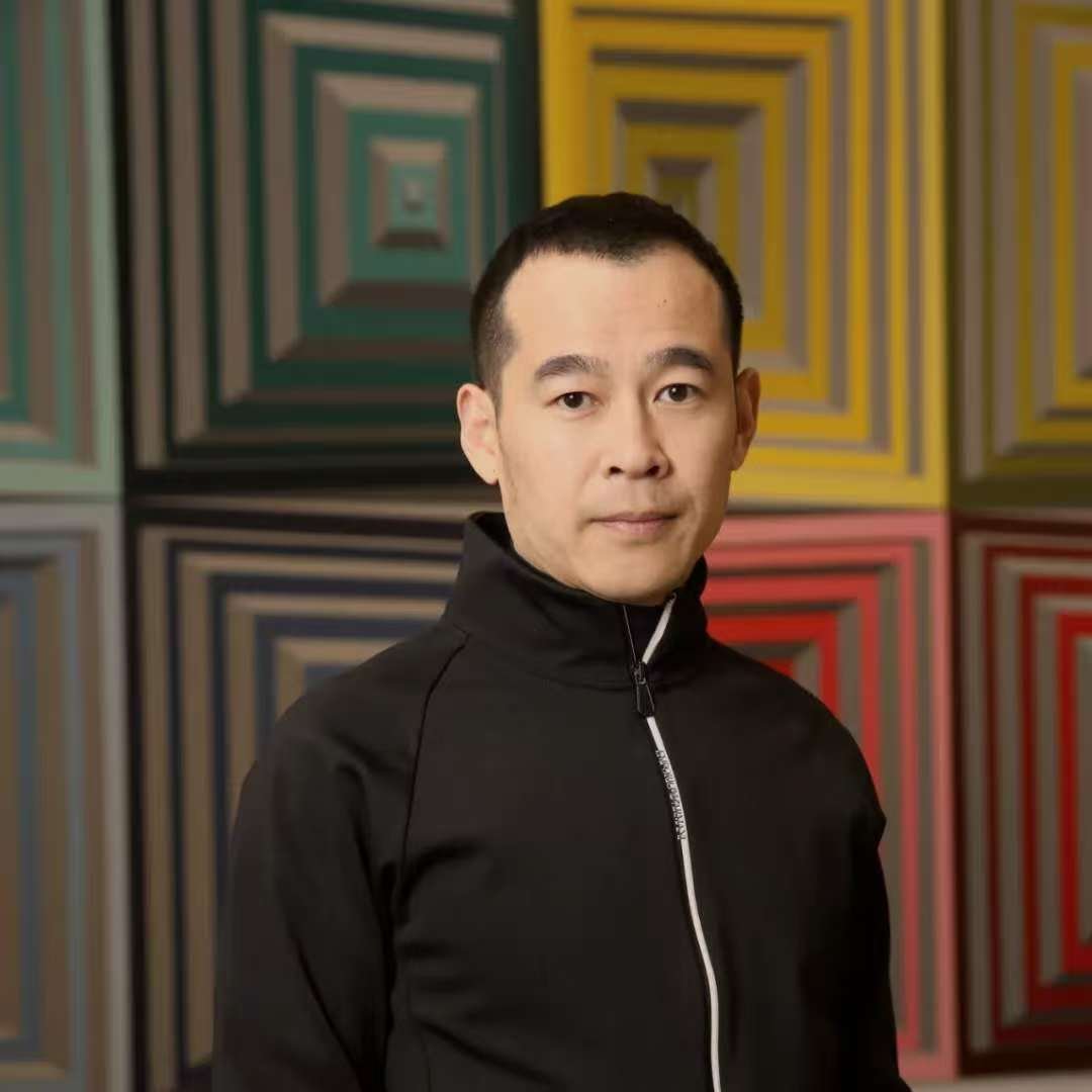A man with short black hair wearing a black zip-up jacket standing in front of a colorful geometric wall art.
