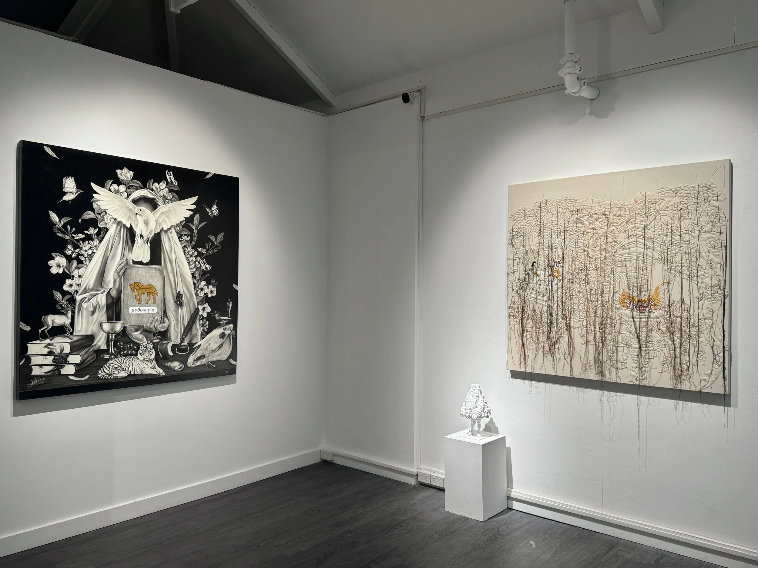 Art gallery with two large artworks on white walls, one black and white with animals and plants, the other in earth tones with detailed trees and mythical creatures, and a white sculpture pedestal in the corner.