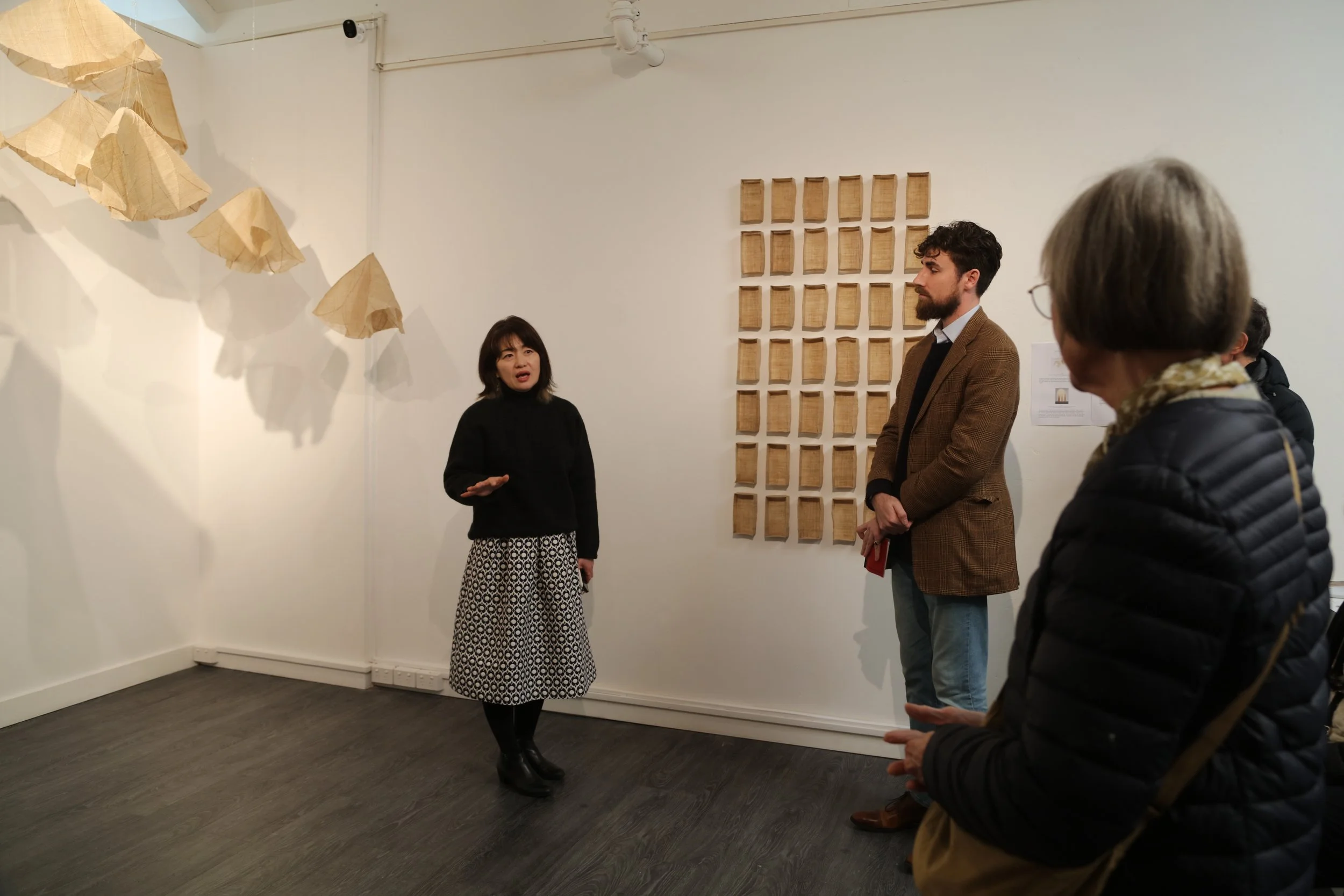 A group of four people are standing in an art gallery. A woman in a black top with a black and white skirt is speaking. Two men and one woman are listening. The background has a grid of 25 wooden panels on the wall and several hanging paper art piece
