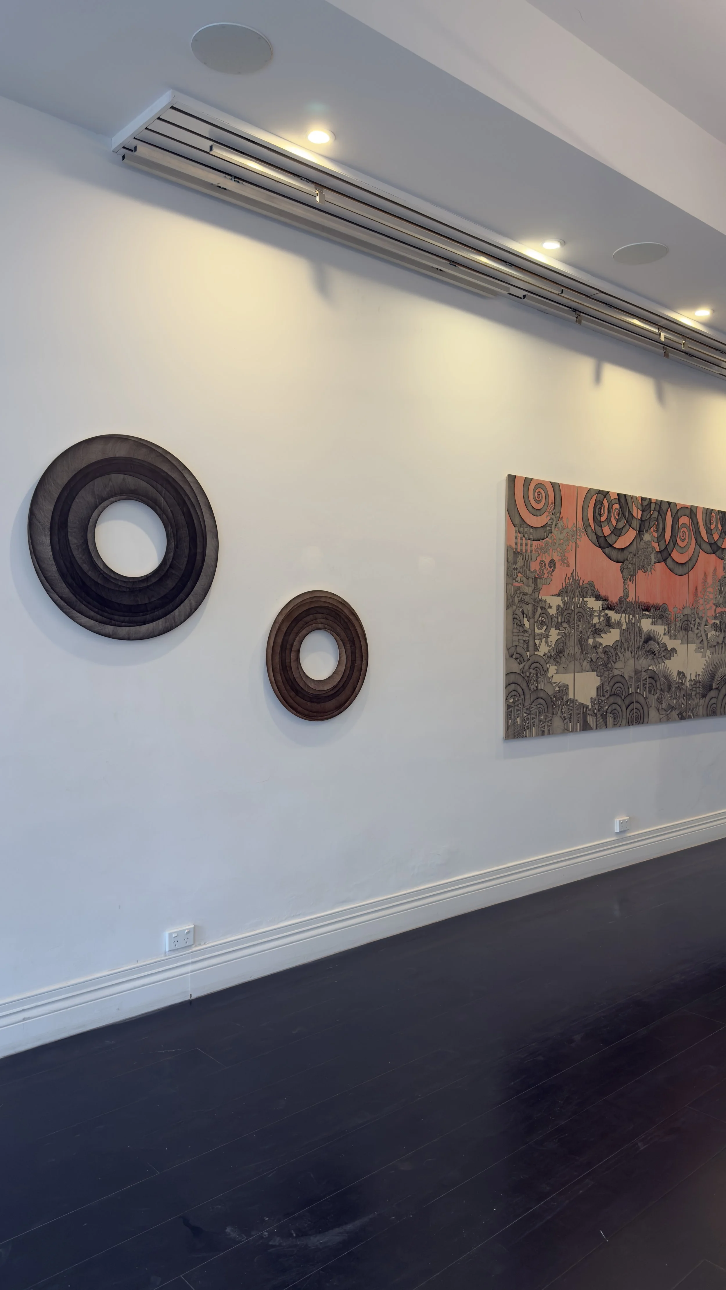 White gallery wall with abstract circular wall art and a larger painting with swirling and detailed scenic elements, above dark hardwood flooring.