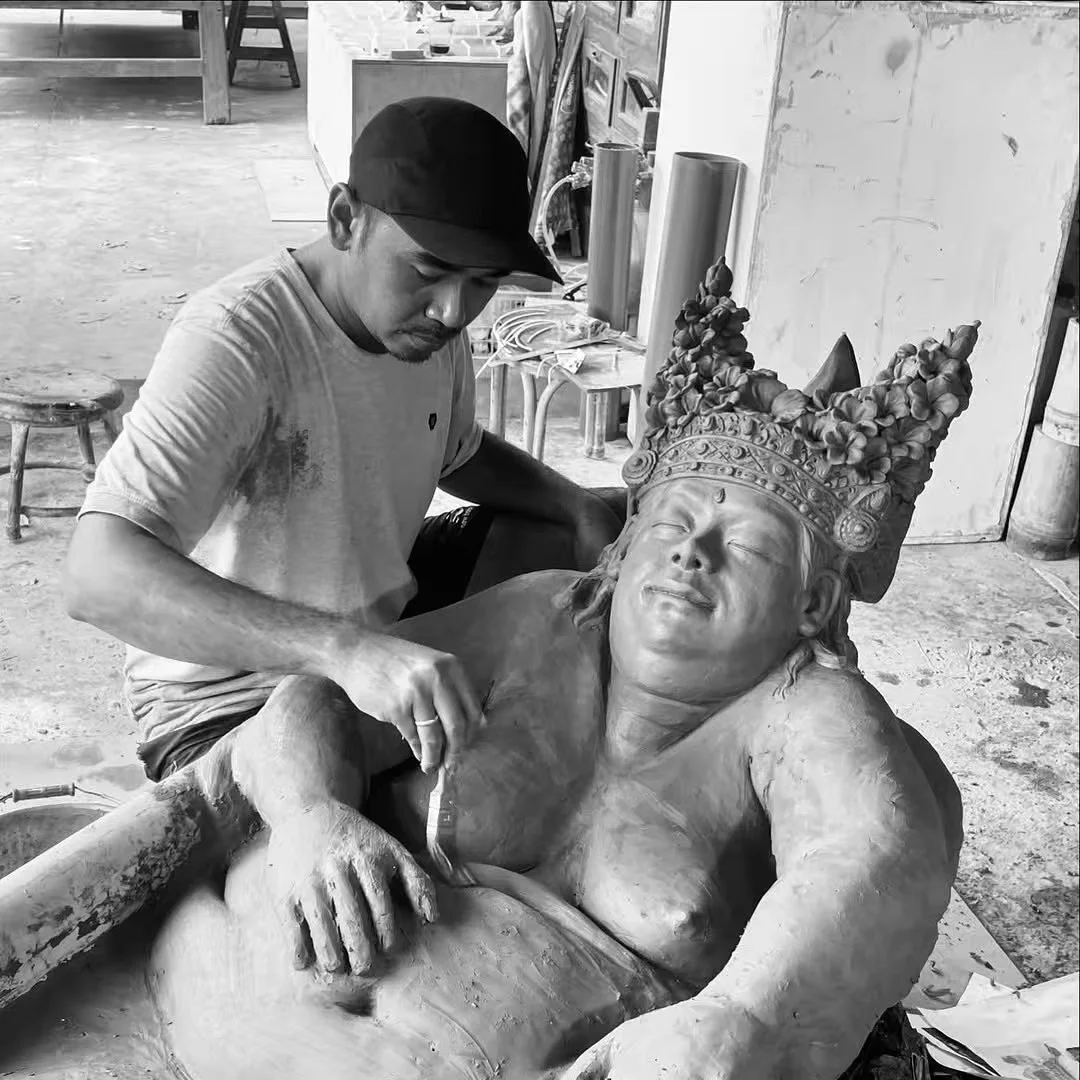 Artist working on a large wooden sculpture. The sculpture depicts a woman with an intricate crown, her eyes closed and a peaceful expression.