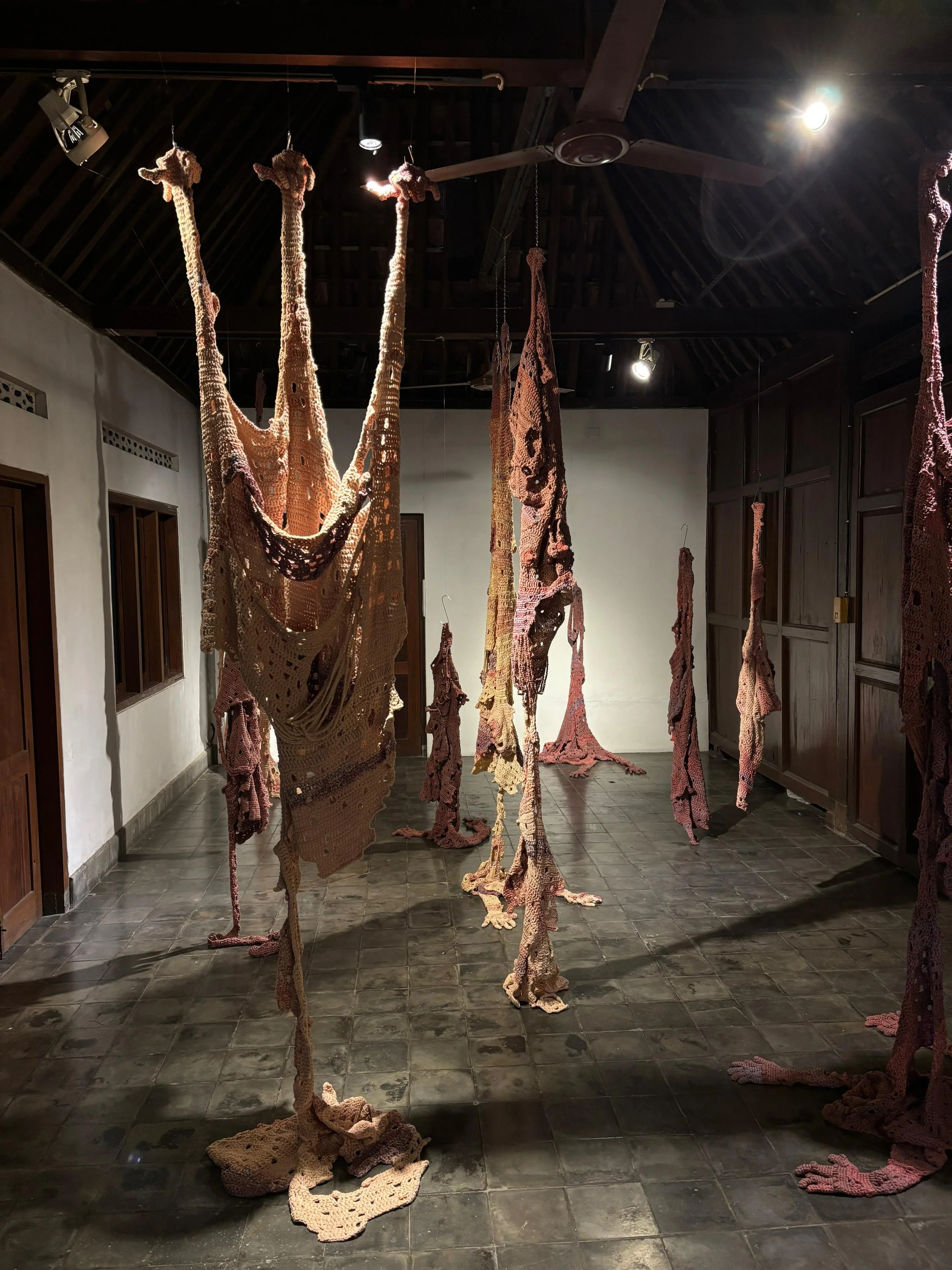 Art installation featuring hanging knitted or crocheted fabric strips resembling giraffe necks and legs in a room with dark tiled floor and wooden walls, illuminated by ceiling lights.