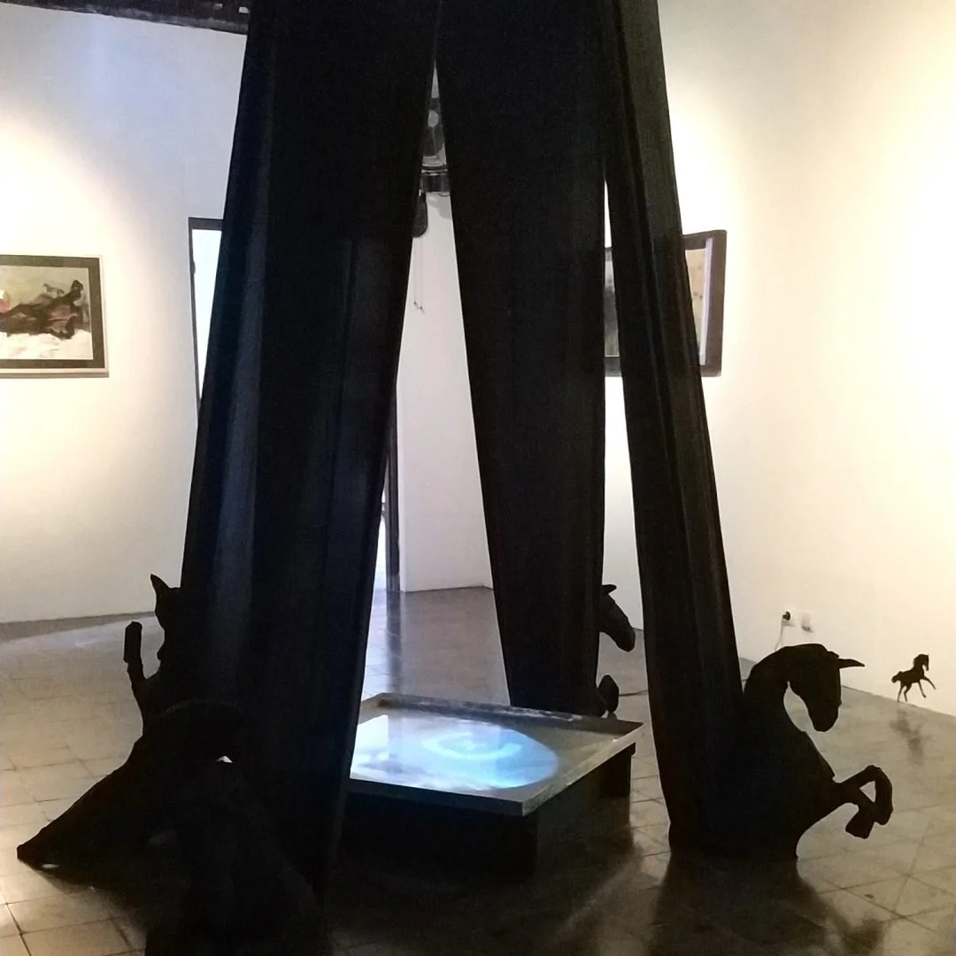 Art installation featuring black horse-shaped silhouettes with a reflective surface and black curtains in a gallery setting.
