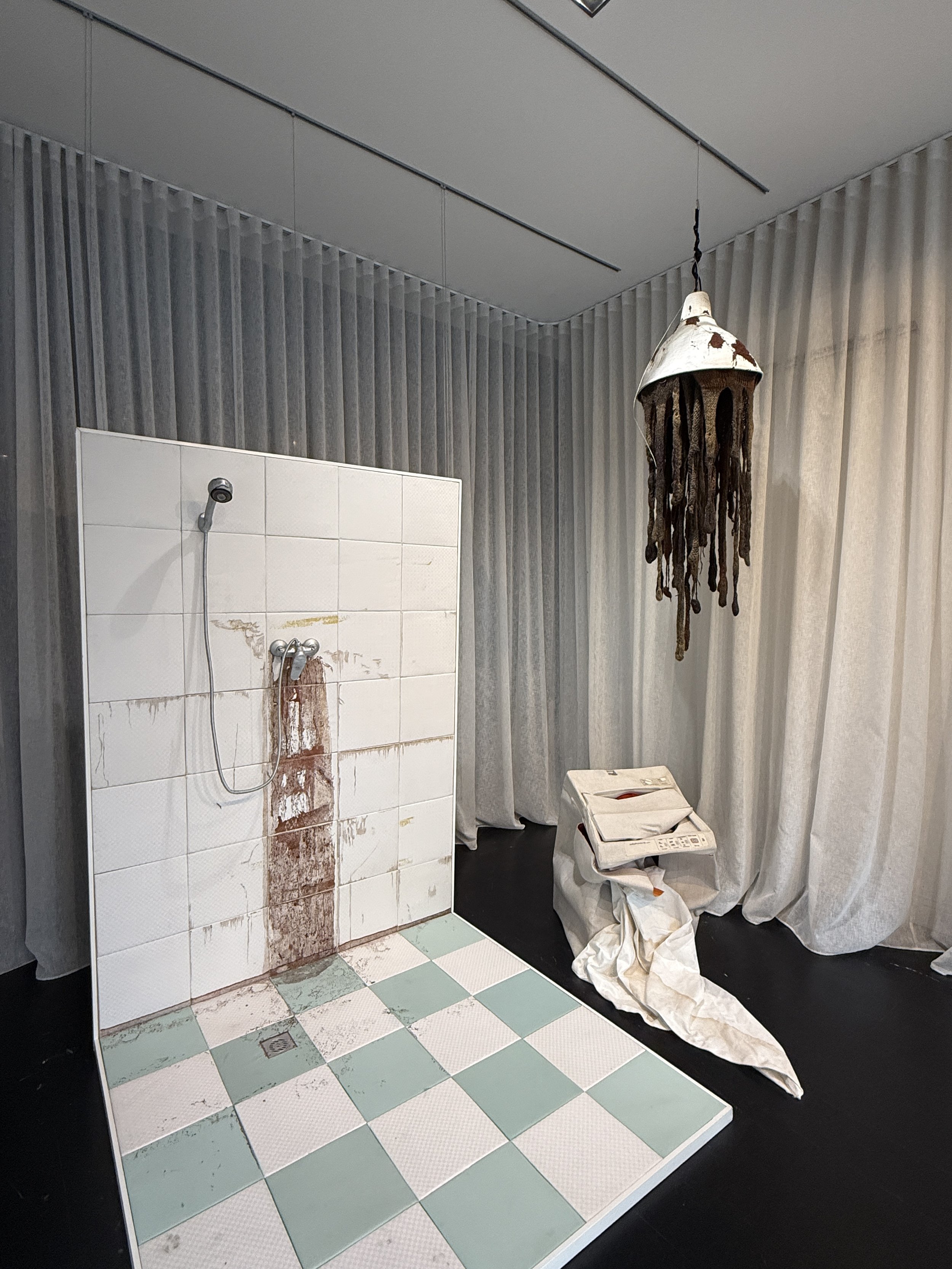 A bathroom scene with a partition of white tiles, some stained or discolored, bordering a shower area. The shower wall has a handheld showerhead attached, with a hose hanging down. The bathroom floor has square tiles in a checkerboard pattern of whit