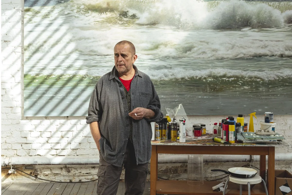 A man standing in front of a brick wall with a large ocean wave painting behind him, a table with spray paint cans, paint bottles, and art supplies, and sunlight casting shadows on the wall.