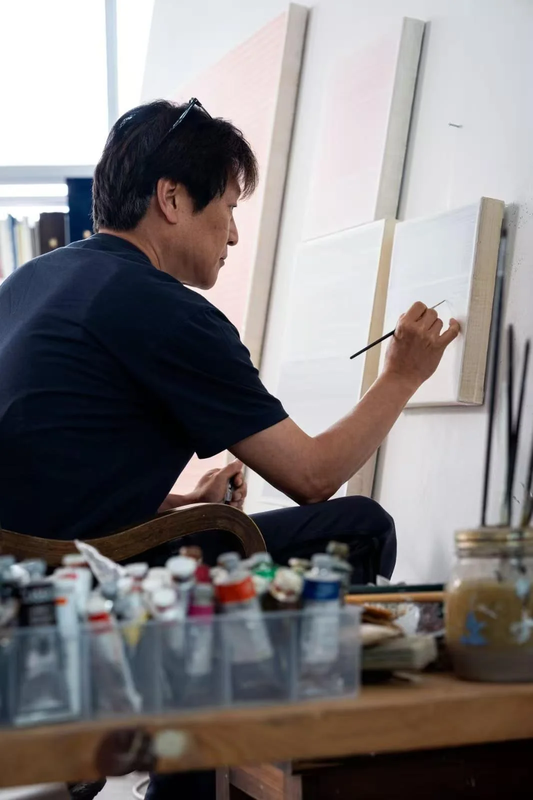 A man seated at a desk painting on a canvas, with art supplies like paint jars and brushes nearby.